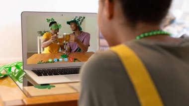 Karışık ırkçı kadın, erkek arkadaşlarına St. Patrick Günü videosu çekiyor. Evde dizüstü bilgisayarla kadeh kaldırıyor. Karantina süresince evde tecrit edilmiş İrlandalı koruyucu azizlerin gününü kutluyoruz..