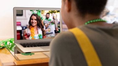 Karışık ırkçı kadın dizüstü bilgisayardaki gülümseyen bayan arkadaşına Aziz Patrick Günü videosu çekiyor. Karantina süresince evde tecrit edilmiş İrlandalı koruyucu azizlerin gününü kutluyoruz..