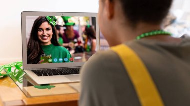 Karışık ırkçı kadın dizüstü bilgisayardaki gülümseyen bayan arkadaşına Aziz Patrick Günü videosu çekiyor. Karantina süresince evde tecrit edilmiş İrlandalı koruyucu azizlerin gününü kutluyoruz..