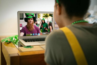 Karışık ırkçı kadın dizüstü bilgisayarında erkek arkadaşına St. Patrick Günü videosu çekiyor. Karantina süresince evde tecrit edilmiş İrlandalı koruyucu azizlerin gününü kutluyoruz..