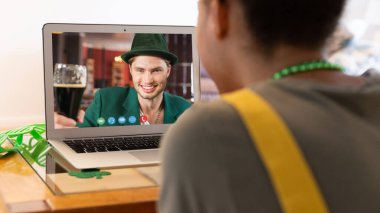 Karışık ırk kadını, Aziz Patrick 'in erkek arkadaşına evde dizüstü bilgisayarında cam kaldırırken video görüşmesi yaptırıyor. Karantina süresince evde tecrit edilmiş İrlandalı koruyucu azizlerin gününü kutluyoruz..