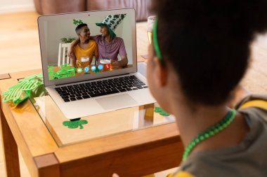 Karışık ırk kadını, dizüstü bilgisayarında iki mutlu erkek arkadaşıyla St. Patrick günü video görüşmesi yapıyor. Karantina süresince evde tecrit edilmiş İrlandalı koruyucu azizlerin gününü kutluyoruz..