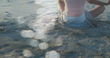 Sahilde yoga yapan ve meditasyon yapan yaşlı beyaz bir kadının üzerinde ışık lekeleri. dünya meditasyon günü ve dijital olarak üretilen video konsepti.