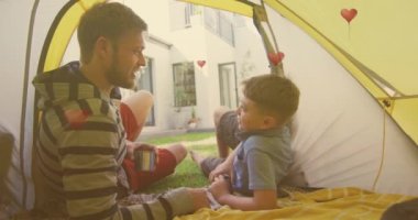 Animation of hearts over caucasian father and son high fiving in tent in garden. school and education during covid 19 pandemic concept digitally generated video.