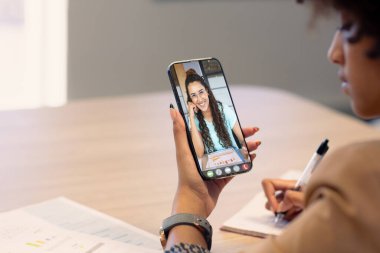 Afrika kökenli Amerikalı iş kadını akıllı telefondan gülümseyen meslektaşını ararken çalışıyor. değiştirilmemiş, ofis, çevrimiçi, teknoloji, video konferansı, tartışma, internet, iş, takım çalışması.