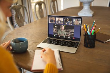 Kafkasyalı iş kadını, beyaz iş arkadaşlarıyla video görüşmesinde çalışırken günlüğe yazı yazıyor. değiştirilmemiş, iş, kablosuz teknoloji, toplantı, çalışma, takım çalışması ve ofis konsepti.