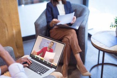 Çift ırklı iş adamı, Afrika kökenli Amerikalı bir erkek meslektaşla video görüşmesi yaparken elinde kağıt tutuyor. değiştirilmemiş, iş, veri, kablosuz teknoloji, toplantı, takım çalışması ve ofis kavramı.