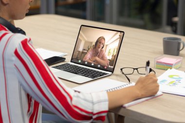 Ofisteki günlüğe yazı yazan Asyalı bir meslektaşla görüntülü görüşme yapan beyaz bir iş kadını. Değiştirilmemiş iş, kablosuz teknoloji, toplantı, çalışma, takım çalışması ve ofis konsepti.