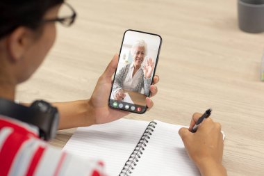 Asyalı iş kadını, kıdemli meslektaşıyla telefonda video konferansı yaparken notlar yazıyor. Günlük, değiştirilmemiş, kablosuz teknoloji, takım çalışması, toplantı, tartışma, iş ve ofis kavramı.