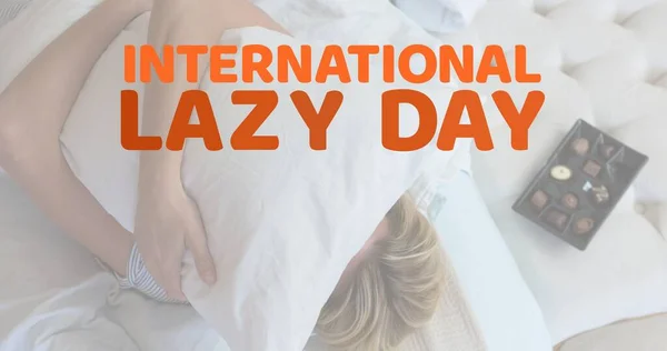 Digital composite image of caucasian woman hugging pillow with international lazy day text at home. Copy space, relaxation, celebrate your inner couch potato, lazy bums, slackers.