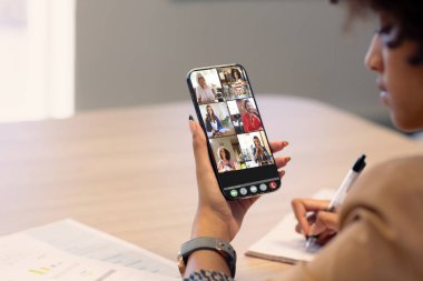 Afrikalı Amerikalı iş kadını cep telefonuyla iş arkadaşlarıyla video konferansı yaparken yazıyor. değiştirilmemiş, kablosuz teknoloji, takım çalışması, toplantı, tartışma, iş, meslek ve ofis.