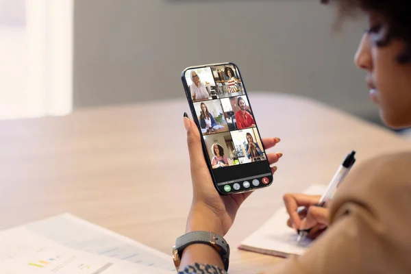 African american businesswoman writing while video conferencing with colleagues over mobile phone. unaltered, wireless technology, teamwork, meeting, discussion, business, occupation and office.