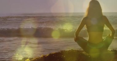 Sahilde yoga yapan beyaz bir kadının arka planda ışık animasyonu. Etkin yaşam tarzı, spor, rahatlama ve deniz konsepti dijital olarak oluşturulmuş video.