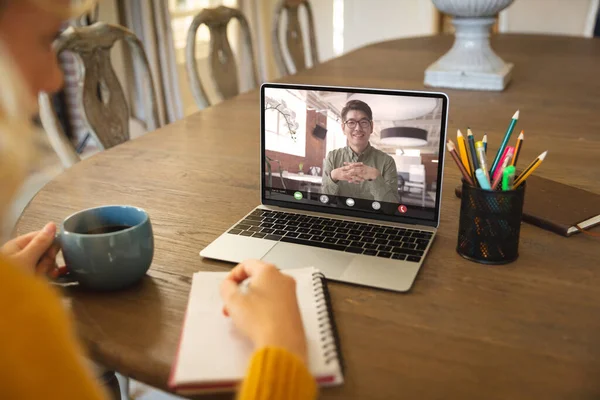 Kafkasyalı iş kadını dizüstü bilgisayarla iş arkadaşıyla video konferansı yaparken not defterine bir şeyler yazıyor. kahve, değiştirilmemiş, kablosuz teknoloji, takım çalışması, toplantı, tartışma, iş ve evden çalışma.