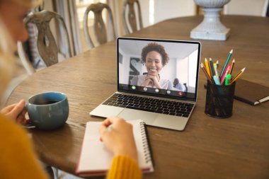 Kafkasyalı iş kadını bilgisayardan iş arkadaşıyla video konferansı yaparken not defterine bir şeyler yazıyor. kahve, değiştirilmemiş, kablosuz teknoloji, takım çalışması, toplantı, tartışma, iş ve evden çalışma.