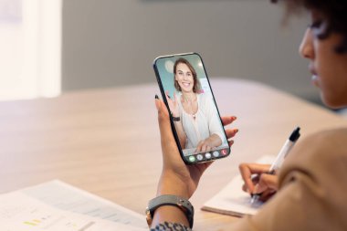 Çift ırklı iş kadını notlar alırken videoda beyaz kadın meslektaşını akıllı telefondan arıyor. değiştirilmemiş, kopyalanmamış, ofis, çevrimiçi, teknoloji, video konferansı, internet, iş, takım çalışması.