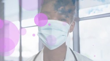 Yüzen pembe noktaların animasyonu mutlu Afrika kökenli Amerikalı kadın doktor maskesinin üzerinde. Coronavirus covid 19 salgını sırasında dijital olarak üretilen ilaç, sağlık ve sağlık hizmetleri.