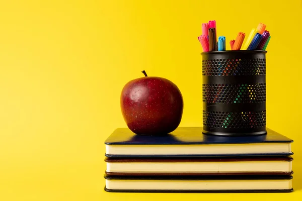 Composition of stack of books, felt tip pens in container and apple on yellow background. School equipment, tools, education and creativity concept.