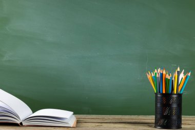 Kara tahtanın üzerindeki tahta masada kitap, defter ve pastel boyaların resmi. Eğitim, öğrenme ve yaratıcılık