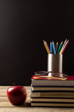 Kitapların, gözlüklerin, kabın içindeki pastel boyaların ve siyah yüzeydeki elmanın dikey görüntüsü. Okul ekipmanları, araçlar, öğrenme ve eğitim kavramı.