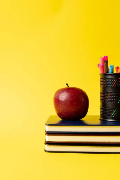 Vertical composition of stack of books, felt tip pens in container and apple on yellow background. School equipment, tools, education and creativity concept.