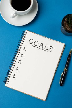 Vertical image of notebook with goals and copy space, coffee and pen on blue surface. Business, working in office, equipment and tools concept.