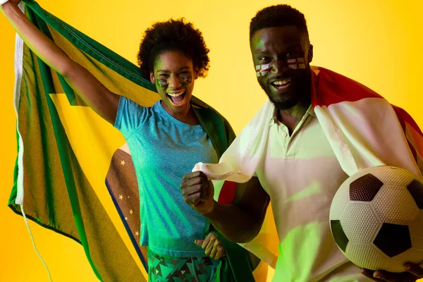 Image of african american soccer fan couple with flags of brazil and england in yellow lightning. Sport, fans, cheering and emotions concept.