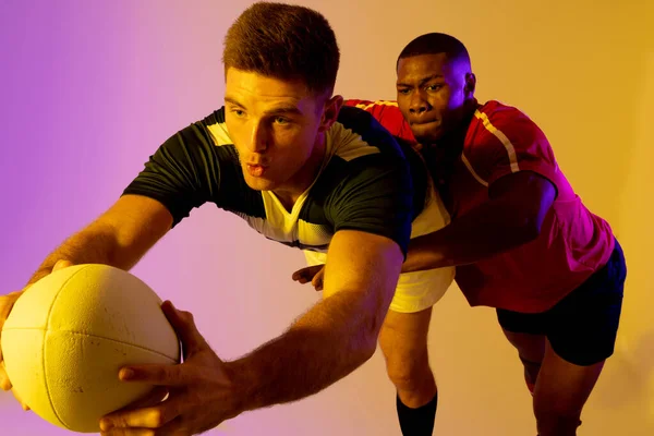 Diverse male rugby players with rugby ball playing over pink lighting. Sport, movement, training and active lifestyle concept.