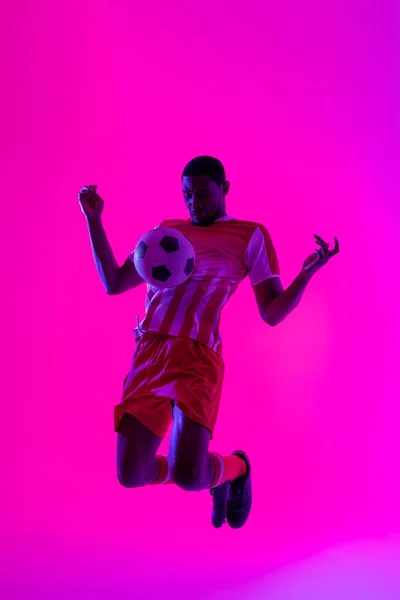 African american male soccer player with football over neon pink lighting. Sport, movement, training and active lifestyle concept.