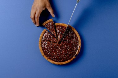 Composition of hands of biracial man cutting pie with knife on blue background. Halloween, autumn, tradition and celebration concept.