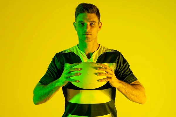 Portrait of caucasian male rugby player with rugby ball over yellow lighting. Sport, movement, training and active lifestyle concept.