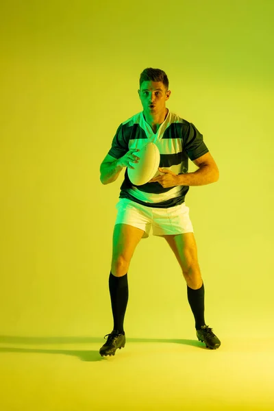 Caucasian male rugby player with rugby ball over yellow lighting. Sport, movement, training and active lifestyle concept.