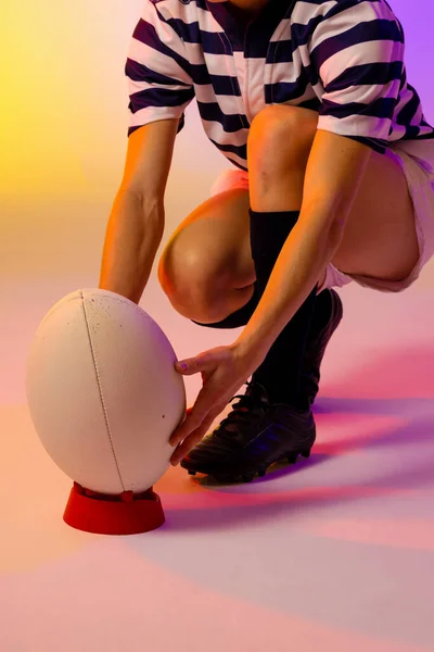 Caucasian female rugby player crouching with rugby ball over neon pink lighting. Sport, movement, training and active lifestyle concept.