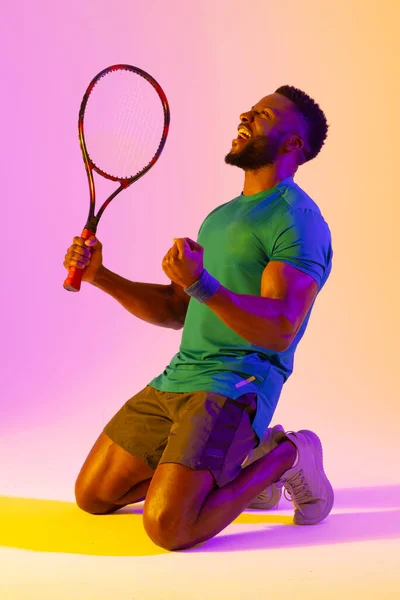 Vertical image of successful african american male tennis player in violet and yellow lighting. Sport, movement, fitness and active lifestyle concept.