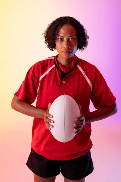 Portrait of african american female rugby player with rugby ball over neon pink lighting. Sport, movement, training and active lifestyle concept.