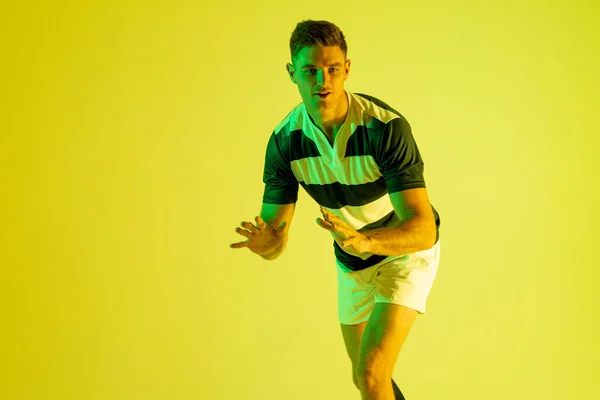 Caucasian male rugby player catching rugby ball over yellow lighting. Sport, movement, training and active lifestyle concept.