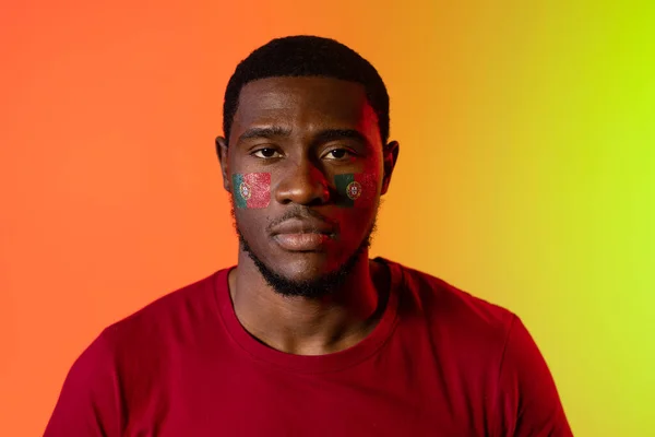 Portrait of african american male supporter with flag of portugal on cheeks over orange lighting. Sport, movement, training and active lifestyle concept.