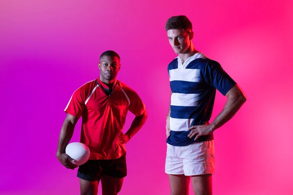 Portrait of diverse male rugby players with rugby ball over pink lighting. Sport, movement, training and active lifestyle concept.