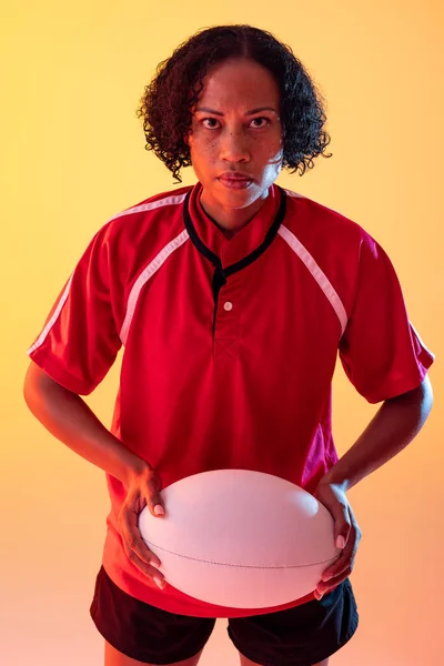 Portrait of african american female rugby player with rugby ball over neon yellow lighting. Sport, movement, training and active lifestyle concept.
