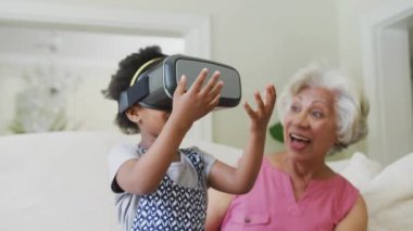 VR kulaklık kullanan Afrikalı Amerikalı büyükanne ve torunun evde oturup mesajlaşması. Dijital olarak oluşturulmuş küresel salgın ve dijital arayüz kavramı.