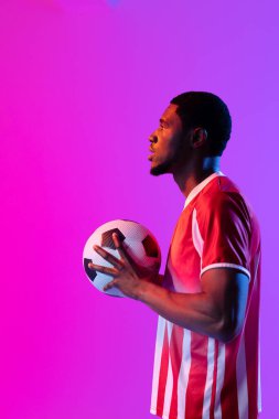 African american male soccer player with football over neon pink lighting. Sport, movement, training and active lifestyle concept.