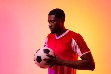 African american male soccer player with football over neon pink lighting. Sport, movement, training and active lifestyle concept.