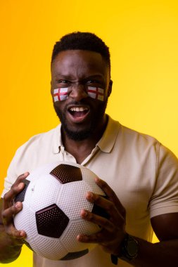Vertical image of african american male fan with flag of england cheering in yellow lighting. Sport, fans, cheering and emotions concept.