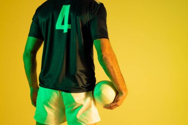 Caucasian male rugby player with rugby ball over yellow lighting. Sport, movement, training and active lifestyle concept.