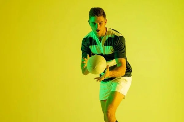 Caucasian male rugby player catching rugby ball over yellow lighting. Sport, movement, training and active lifestyle concept.
