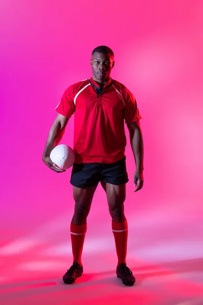 Portrait of african american male rugby player with rugby ball over pink lighting. Sport, movement, training and active lifestyle concept.