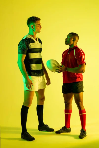 Diverse male rugby players with rugby ball over yellow lighting. Sport, movement, training and active lifestyle concept.