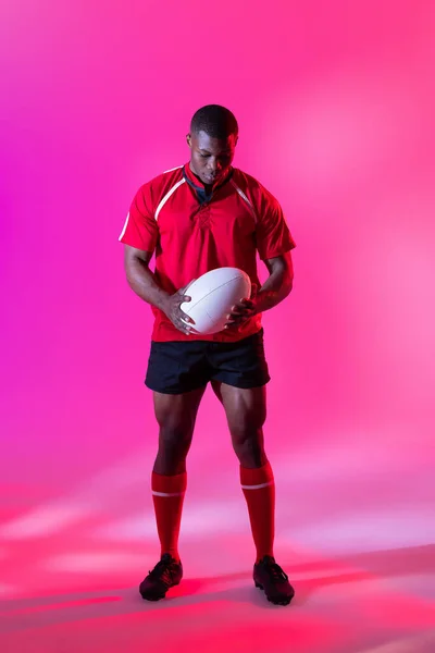 African american male rugby player with rugby ball over pink lighting. Sport, movement, training and active lifestyle concept.