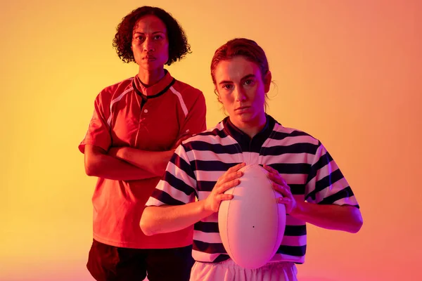 Portrait of diverse female rugby players with rugby ball over neon pink lighting. Sport, movement, training and active lifestyle concept.