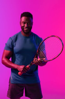 Mor ve pembe neon ışıklı mutlu Afrikalı Amerikalı erkek tenisçinin dikey görüntüsü. Spor, hareket, fitness ve aktif yaşam tarzı konsepti.